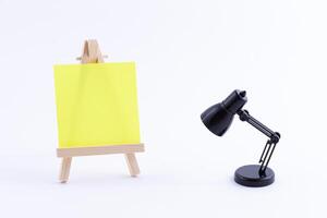 Wooden Easel Miniature with Blank Colored Square Canvas or Memo Paper - Mockup. Mini Wooden Stand with Clean Artboard and Small Black Table Lamp on White Background, Copy Space photo
