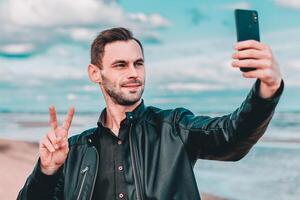 Young Blogger Making Selfie or Streaming at the Beach Using Black Smartphone. Handsome Man in Black Clothes Making Photo Against the Sea at Cloudy Day
