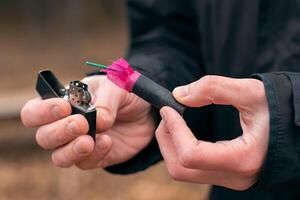 Setting Fire to the Firecracker. Man in Black Clothes Lighting Up the Petard. Firing Up the Pyrotechnics with a Black Gasoline Lighter Outdoors photo