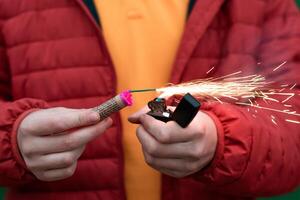 Man in Red Jacked Lighting Up Firecracker in his Hand Using Gasoline Lighter. Guy Getting Ready for New Year Fun with Fireworks or Pyrotechnic Products photo