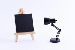 Wooden Easel Miniature with Blank Black Square Canvas for Artists and Painters - Mockup. Mini Wooden Stand with Clean Artboard and Small Black Table Lamp on White Background, Copy Space photo