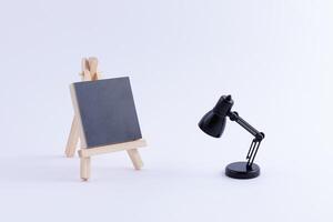 Wooden Easel Miniature with Blank Black Square Canvas for Artists and Painters - Mockup. Mini Wooden Stand with Clean Artboard and Small Black Table Lamp on White Background, Copy Space photo
