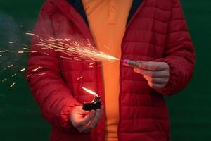 Man in Red Jacked Lighting Up Firecracker in his Hand Using Gasoline Lighter. Guy Getting Ready for New Year Fun with Fireworks or Pyrotechnic Products photo