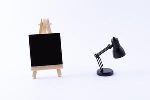 Wooden Easel Miniature with Blank Black Square Canvas for Artists and Painters - Mockup. Mini Wooden Stand with Clean Artboard and Small Black Table Lamp on White Background, Copy Space photo