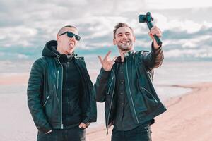 Two Handsome Smiling Friends Making Selfie Using Action Camera with Gimbal Stabilizer at the Beach. Youthful Men in Black Clothes Having Fun by Making Photos