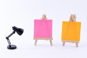 Two Wooden Easels Miniature with Blank Colored Square Canvas or Memo Paper - Mockup. Mini Wooden Stand with Clean Artboard and Small Black Table Lamp on White Background, Copy Space photo