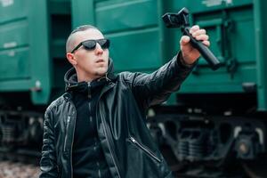 Handsome Guy in Black Clothes and Sunglasses Making Selfie or Streaming Using Action Camera with Gimbal Camera Stabilizer at Railway photo