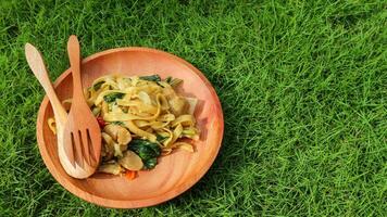 Kwetiaw or Kwetiau is Chinese food which is popular in Indonesia, cooked by frying added with chicken, seafood and complementary vegetables. Served in wooden plate on green grass background. photo
