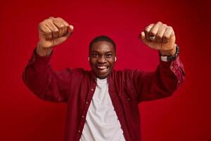 joven africano americano hombre se regocija con manos a cámara, aislado en rojo antecedentes estudio retrato foto