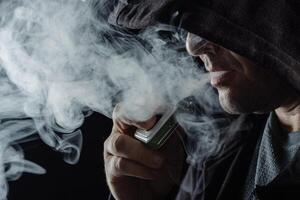 Man with vaping mod exhaling steam at black studio background. Bearded guy smoking e-cigarette to quit tobacco photo
