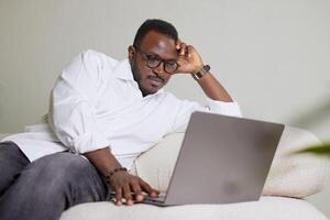 Handsome Black African American Man Working on Laptop Computer while Sitting on a Sofa in Cozy Living Room. Freelancer Working From Home. Browsing Internet, Using Social Networks, Having Fun in Flat. photo