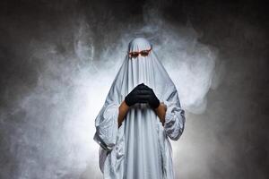 man in scary ghost costume in smoke on grey mysterious background in studio. Minimal halloween ghost concept. photo