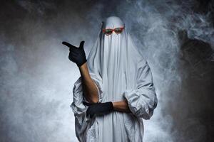 Man in ghost costume pointing at something on gray background in smoke with space for text. Halloween celebration photo