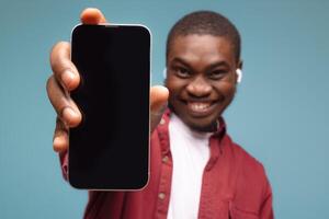 Mobile advertisement. Overjoyed black man pointing at cellphone with empty white screen on blue studio background, mockup for app or website. Cellphone display template photo