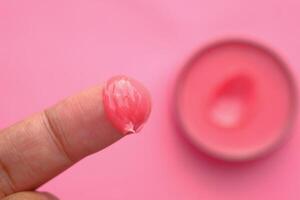 A Detailed CloseUp Image Showing Pink Cream on a Finger, with a Container in the Background photo