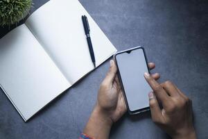 A person is using a smartphone while sitting next to a notebook and a pen on a dark surface photo