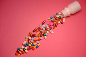 A collection of colorful pills and capsules spilling out from an open bottle onto a pink background photo