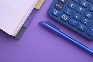 A Calculator, Notebook, and Pen placed on a vibrant Purple Background ideal for Study or Office use photo