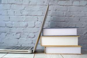 A Laptop and a Stack of Books are resting on a Wooden Table against a Brick Background photo