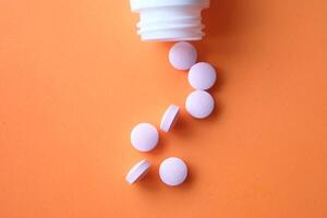 Colorful pills spill from an open white bottle onto a vibrant orange background photo