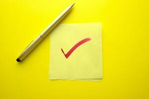 A Checkmark on a Sticky Note with a Pen resting beside it on a Bright Yellow Background photo