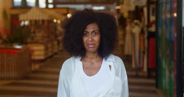 African American woman with shaggy hairstyle, wearing white shirt, expressing shock, dissatisfaction, disagreement and gesturing looking at camera, showing thumb down over blurred store interior video