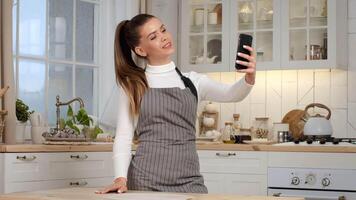 A young woman stands in a bright kitchen, smiling as she takes a selfie with her phone while wearing an apron. video