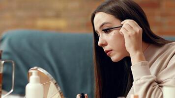 A young woman sits at a table, focused on applying mascara in front of a mirror. The room is warmly lit, creating a comfortable atmosphere while she perfects her makeup look. video