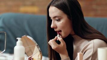 A young woman is focused on applying lipstick in front of a mirror. The warm, inviting ambiance features a soft couch and various beauty products on a table. video