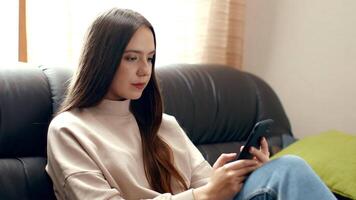 A young woman sits comfortably on a couch, focused on her smartphone. Soft natural light filters through a window, creating a warm and inviting atmosphere in her living space. video