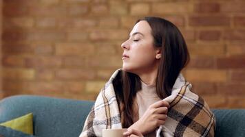 A young woman, cozy in a blanket, thoughtfully sips her warm drink while sitting on a couch. The setting features a backdrop of textured brick walls, enhancing the comforting atmosphere. video