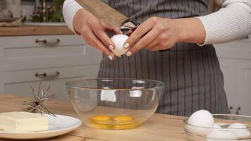 A person in a striped apron cracks an egg into a bowl while holding a knife in a modern kitchen setting. video