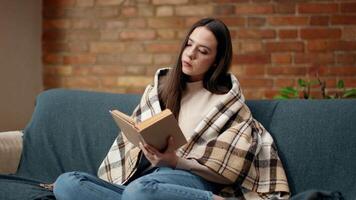 A woman sits comfortably on a couch, wrapped in a warm blanket, deeply engrossed in reading a book amidst a charming, cozy living space featuring a brick wall. video