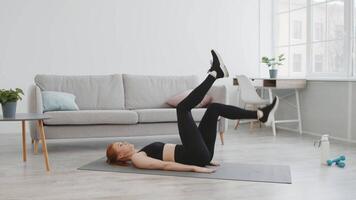 A woman in workout attire performs leg raises on a yoga mat in a bright, minimalist room. Natural light fills the space, creating an inviting atmosphere for her fitness routine. video