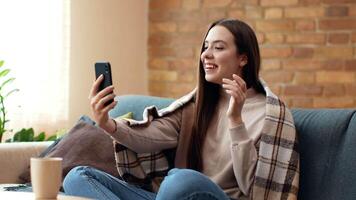 A young woman sits comfortably on a couch, wrapped in a blanket, smiling as she engages in a call on her phone. Sunlight streams through the window, adding warmth to the room. video