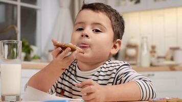 A child relishes a cookie with milk, displaying a playful expression while sitting at a kitchen table filled with colorful treats. video