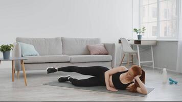 A woman exercises on a yoga mat, doing side leg raises in a bright living room with a cozy sofa. The space features large windows, a modern desk, and indoor plants, emphasizing a healthy lifestyle. video