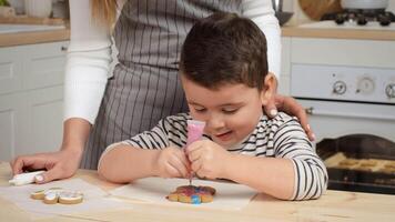 ein Kind gilt bunt Glasur zu ein Lebkuchen Plätzchen während ein Erwachsene bietet an Orientierungshilfe im ein hell, einladend Küche. video