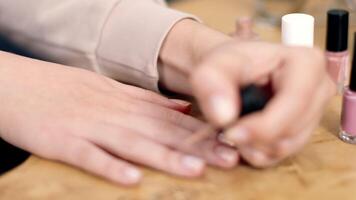 A woman is carefully applying light pink nail polish to her fingernails while seated at a wooden table. Various nail polish bottles are visible in the background, creating a calming atmosphere. video