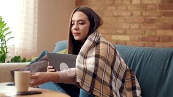 A woman sits on a couch in a cozy living room, focused on her laptop while wrapped in a warm blanket, surrounded by soft lighting and a relaxed atmosphere. video