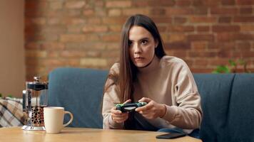 A young woman sits on a sofa, focused on playing a game. She holds a gaming controller, with a coffee mug nearby. The warm atmosphere suggests a relaxing afternoon. video