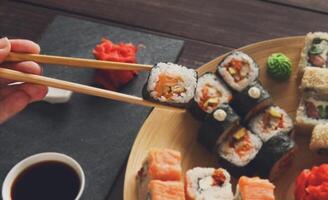 Japanese food restaurant, sushi maki roll plate, platter set. Hand take roll. Set for two with chopsticks, ginger, soy, wasabi. Sushi at rustic wood background and black stone. POV at black. Closeup. photo