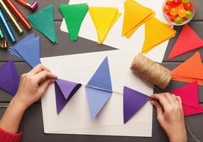Birthday handmade background. Female hands making party paper decorations and colorful flags garland. DIY accessories on gray wooden planks, top view. Holiday organization, hobb, handicraft concept photo