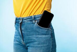 Unrecognizable woman having smartphone with empty screen in her jeans pocket on blue studio background, mockup for mobile app or website design. Space for your ad template photo