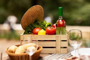 A wooden crate filled with fresh tomatoes, lettuce, and a green wine bottle sits on a table outdoors. Surrounding the crate are glasses and a basket of bread, perfect for a picnic. photo