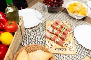 A rustic wooden table is set for a casual summer gathering featuring an array of fresh foods like skewers, fruits, and wine. The atmosphere is inviting and relaxed, perfect for dining. photo