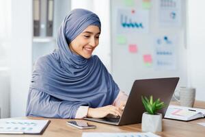 Office Job. Happy islamic woman in hijab working on laptop at desk, typing on keyboard and looking at computer screen. photo