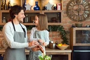 Joyful Young Dad And Little Daughter Cooking Vegetable Salad For Lunch Together, Adding Seasoning To Bowl, Enjoying Healthy Homemade Food, Having Fun At Home, Wearing Aprons And Smiling, Free Space photo