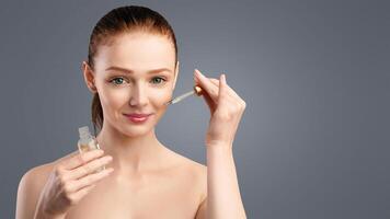 Skincare. Smiling Redhaired Woman Applying Cosmetic Oil On Face Caring For Facial Skin Posing Standing On White Background In Studio, Looking At Camera. Spa And Beauty Concept photo