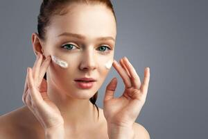 Skincare. Ginger-Haired Young Lady Applying Facial Cream Moisturizing And Caring For Skin Standing Posing Over Gray Studio Background. Skin Care Cosmetics For Youth Concept photo